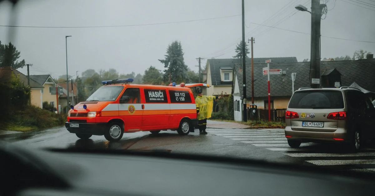 A fire department vehicle blocks a street intersection during wet weather, guided by a uniformed official.