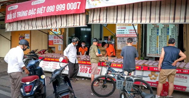 People buying lottery tickets from street vendors. Motorcycles parked nearby on the sidewalk.