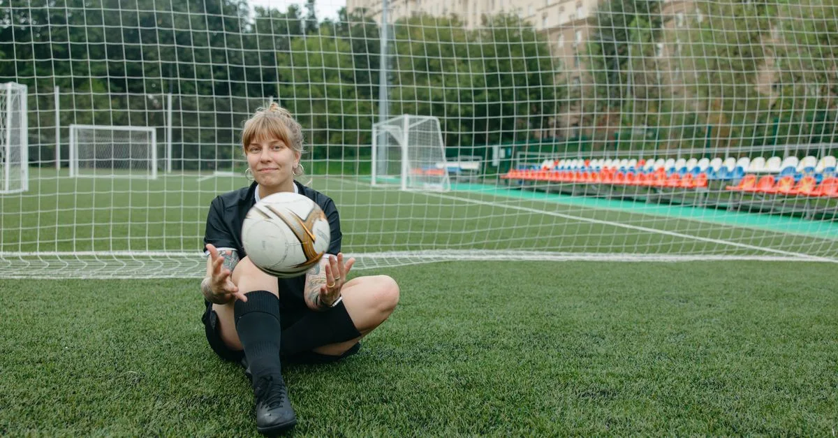 A female soccer player sitting with a ball on a soccer field, enjoying a break.