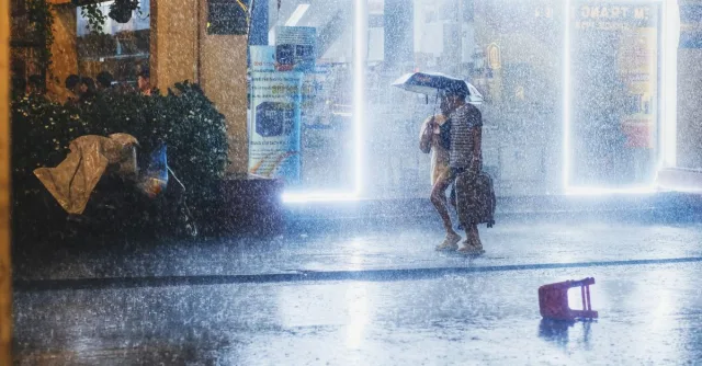 A nighttime urban scene with heavy rain, people under umbrellas, and brightly lit storefronts.