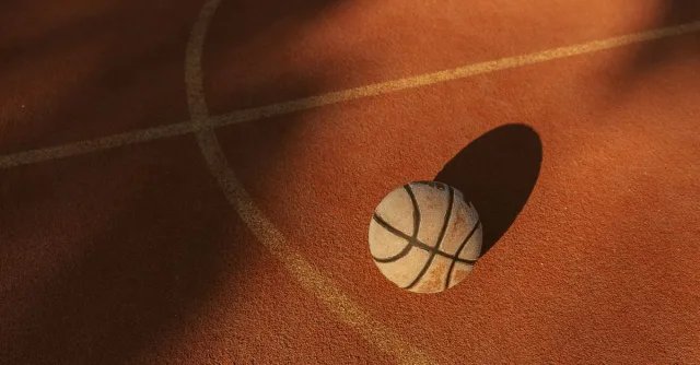 A basketball sits alone under sunlight creating bold shadows on an outdoor court.
