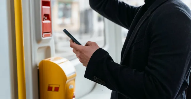 Adult in a black coat checking smartphone while commuting on public transport.