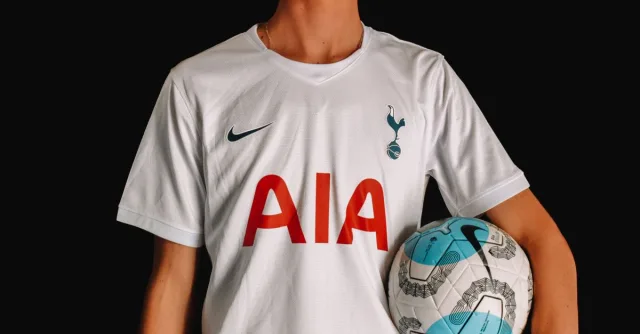 Adult soccer player in Tottenham Hotspur jersey holding a ball against a dark background.