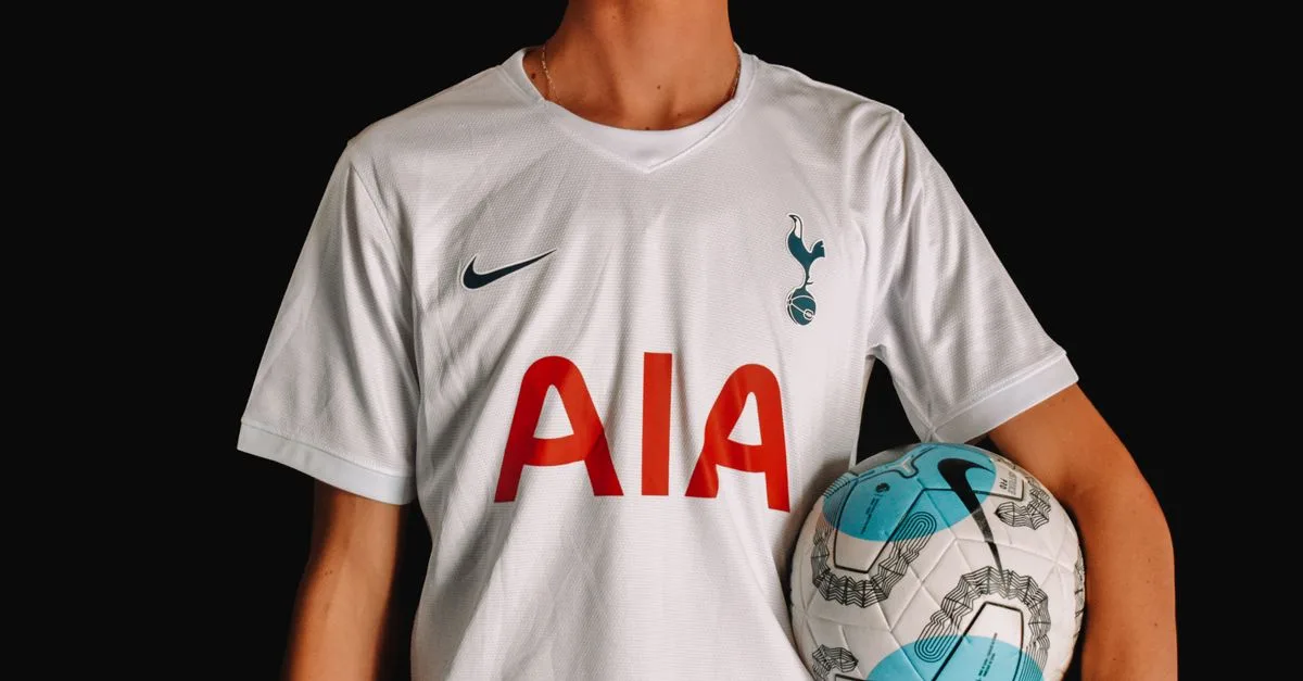Adult soccer player in Tottenham Hotspur jersey holding a ball against a dark background.