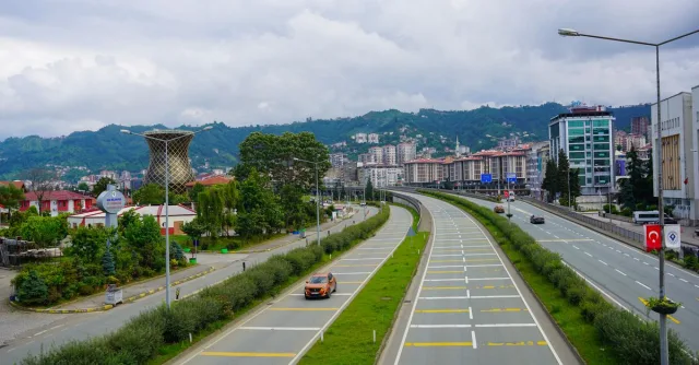 A vibrant view of a road in Rize, Türkiye, with mountains and cityscape under a cloudy sky.