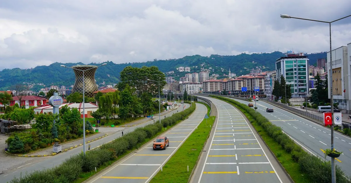 A vibrant view of a road in Rize, Türkiye, with mountains and cityscape under a cloudy sky.