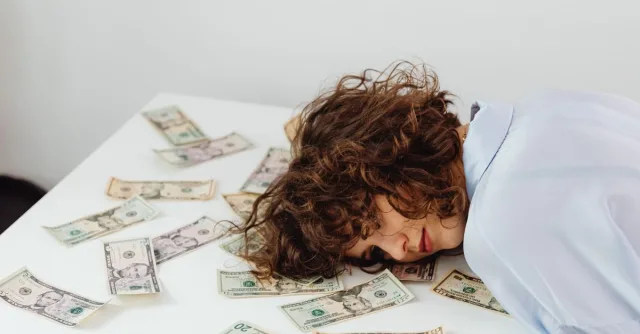 A woman with curly hair resting her head on a table covered in US dollar bills.