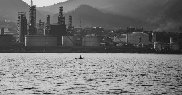 A lone kayaker paddles on a lake with an oil refinery in the backdrop of mountains.