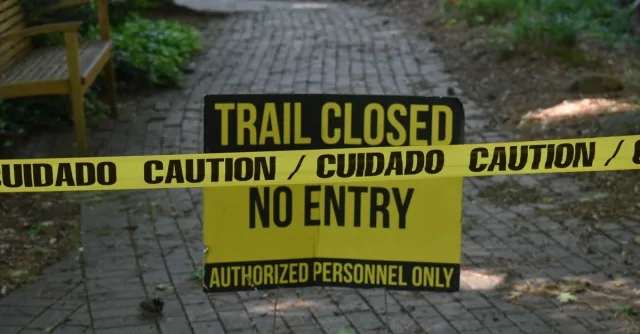 Trail closed sign with caution tape blocking pathway in a park setting.