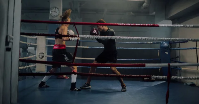 A man and woman spar in an indoor boxing ring, showcasing strength and athleticism.