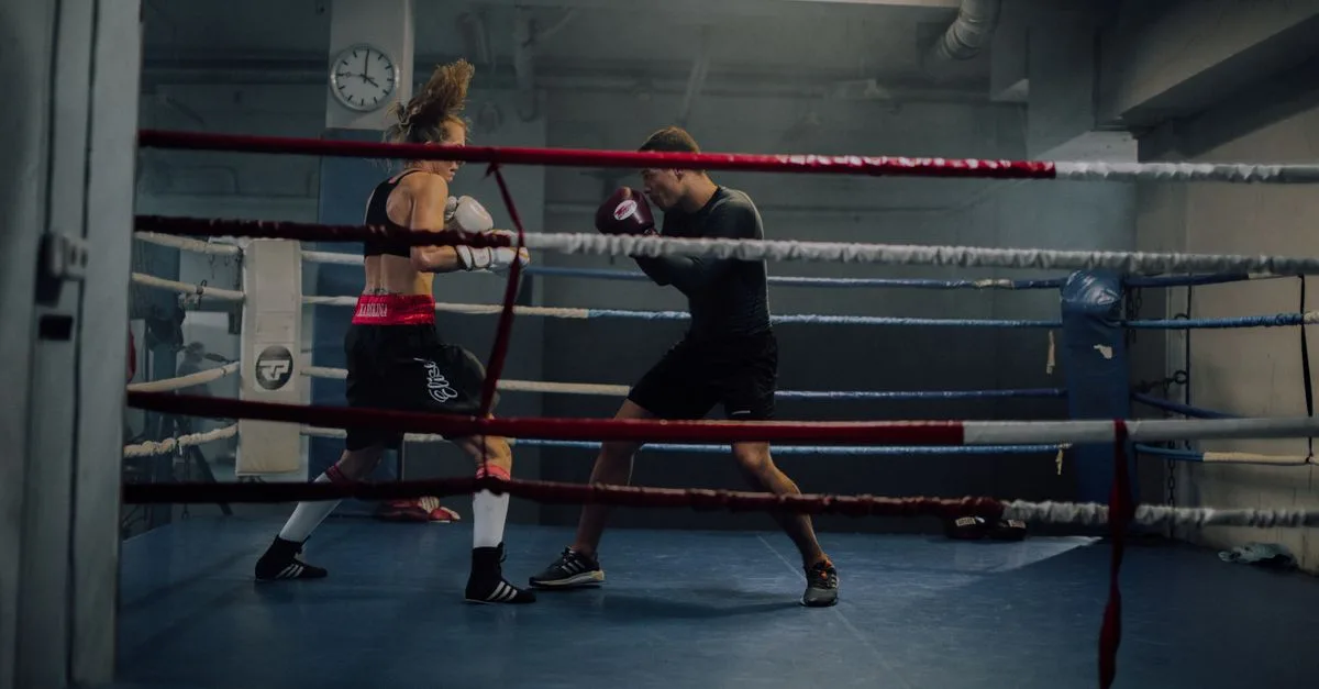 A man and woman spar in an indoor boxing ring, showcasing strength and athleticism.