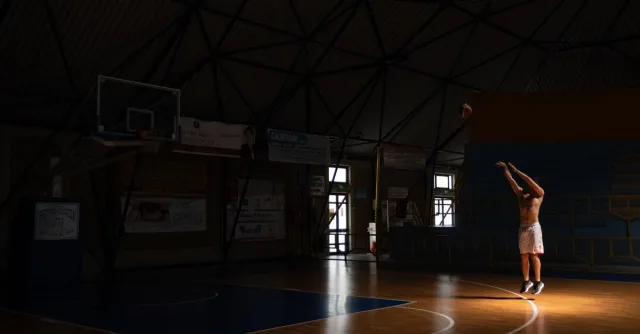 A man practices basketball shots in a dimly lit indoor court, creating a dramatic and focused atmosphere.
