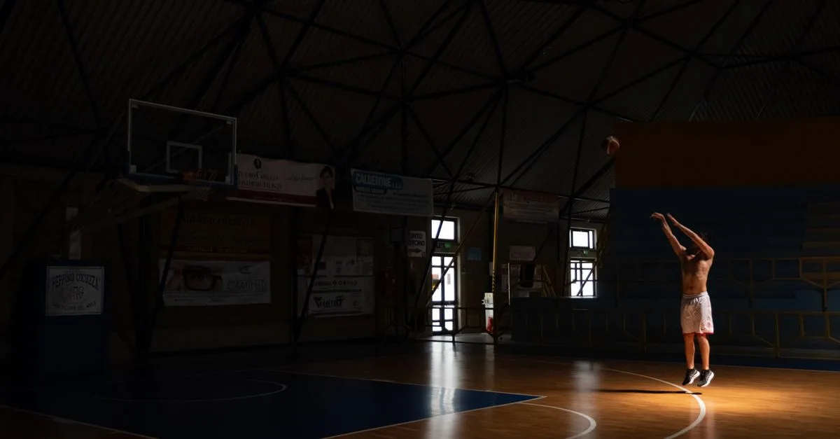A man practices basketball shots in a dimly lit indoor court, creating a dramatic and focused atmosphere.