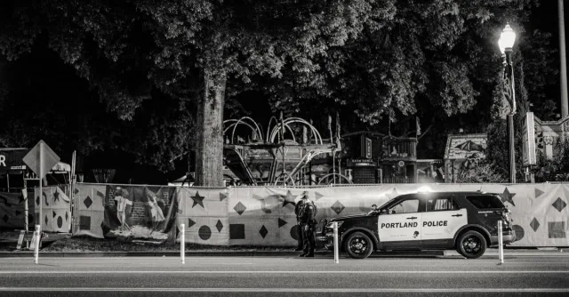 Black and white nighttime street view featuring a Portland police car parked by a decorated fence.
