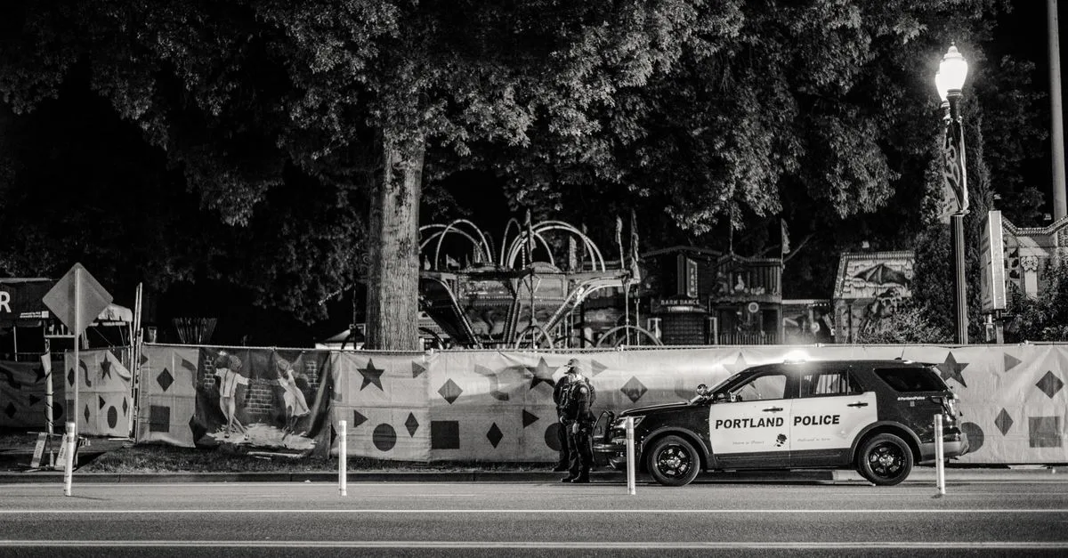 Black and white nighttime street view featuring a Portland police car parked by a decorated fence.