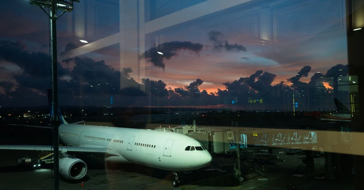 Through glass modern white plane connected to airport terminal with airbridge and ready for boarding against spectacular cloudy purple sky in late evening