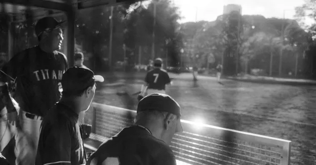 Baseball players in a dugout during a game in a sunlit Tokyo park.