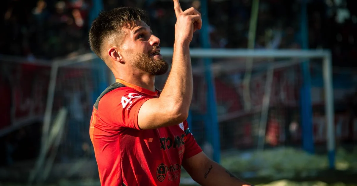 A football player in a red jersey celebrating with a finger point during a match outdoors.
