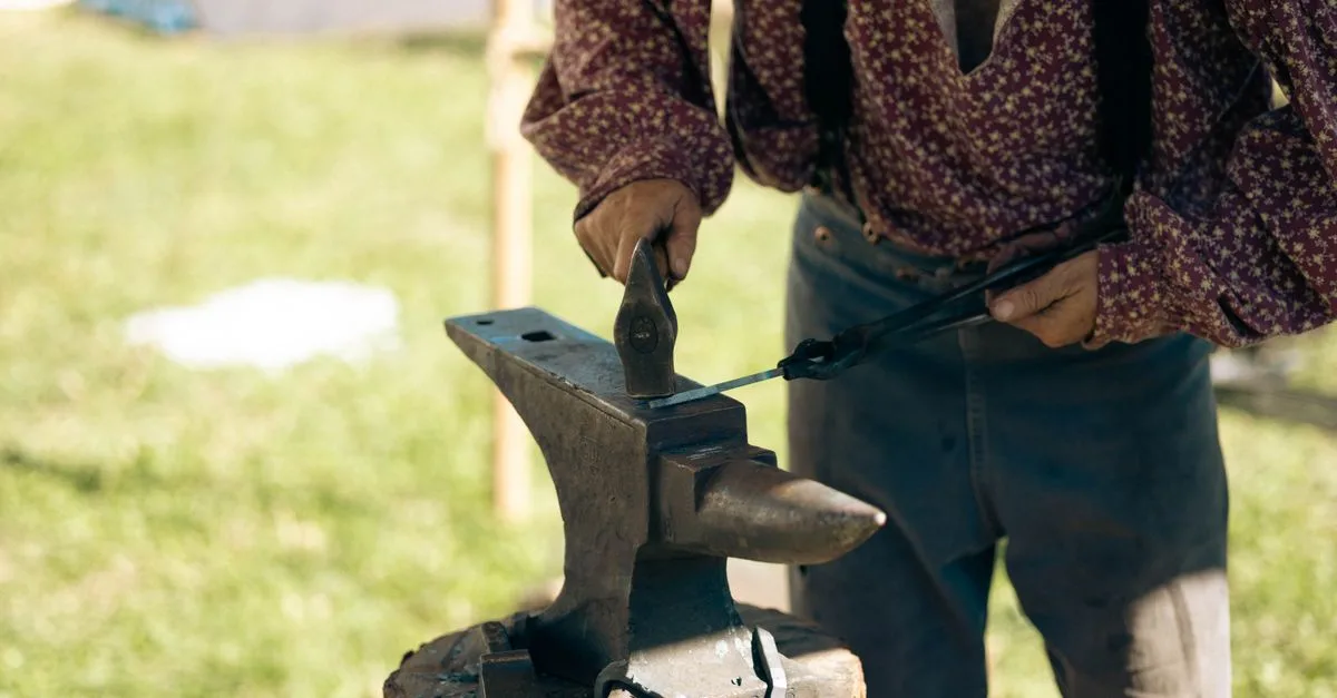 A blacksmith in traditional attire uses a hammer and anvil outdoors, showcasing artisan skills.