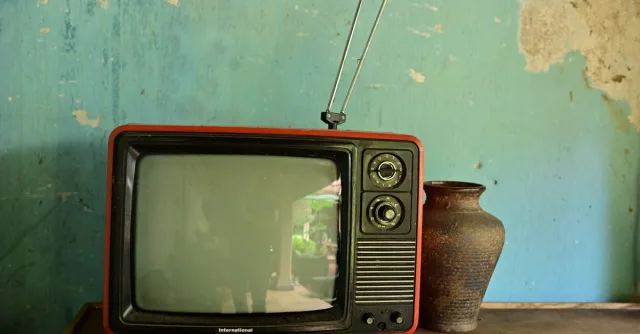 A vintage television with antenna beside a ceramic vase on a worn wooden surface against a rustic wall.