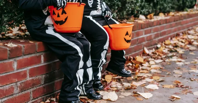 Crop anonymous children dressed as scary skeletons sitting on red brick fence with jack o lantern buckets on Halloween day