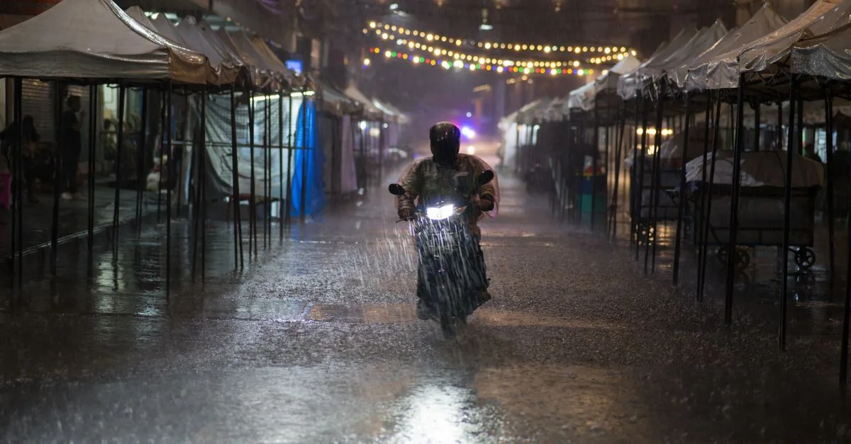 Motorcycle rider braves heavy rain in Bangkok street lined with tents and string lights at night.