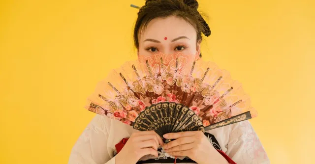 Asian woman in traditional hanfu holding a decorative fan against a yellow backdrop.