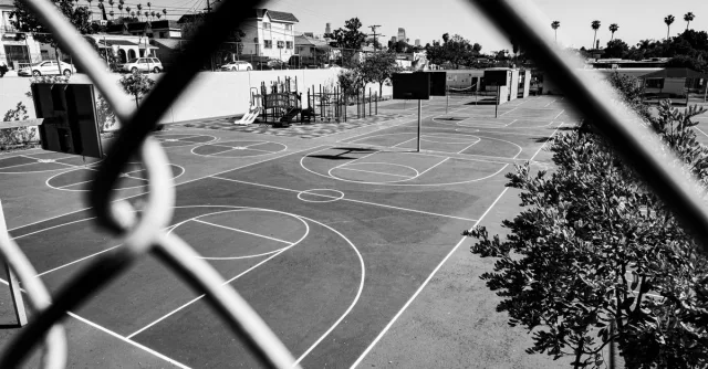 Black and white image of an outdoor basketball court viewed through a chain-link fence.