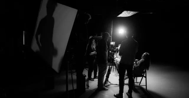 A black and white view of a film crew working on set with dramatic lighting.