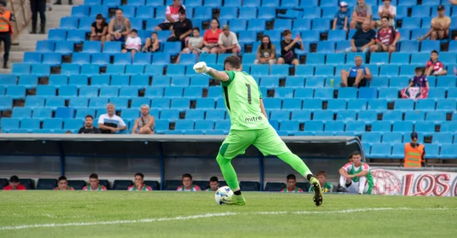 Goalkeeper in green jersey kicking the ball during a soccer match with spectators in a stadium.